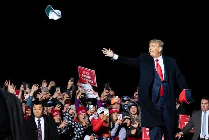 The president, who is maskless tosses a MAGA hat into the crowd; a section of the crowd behind him appears mostly maskless